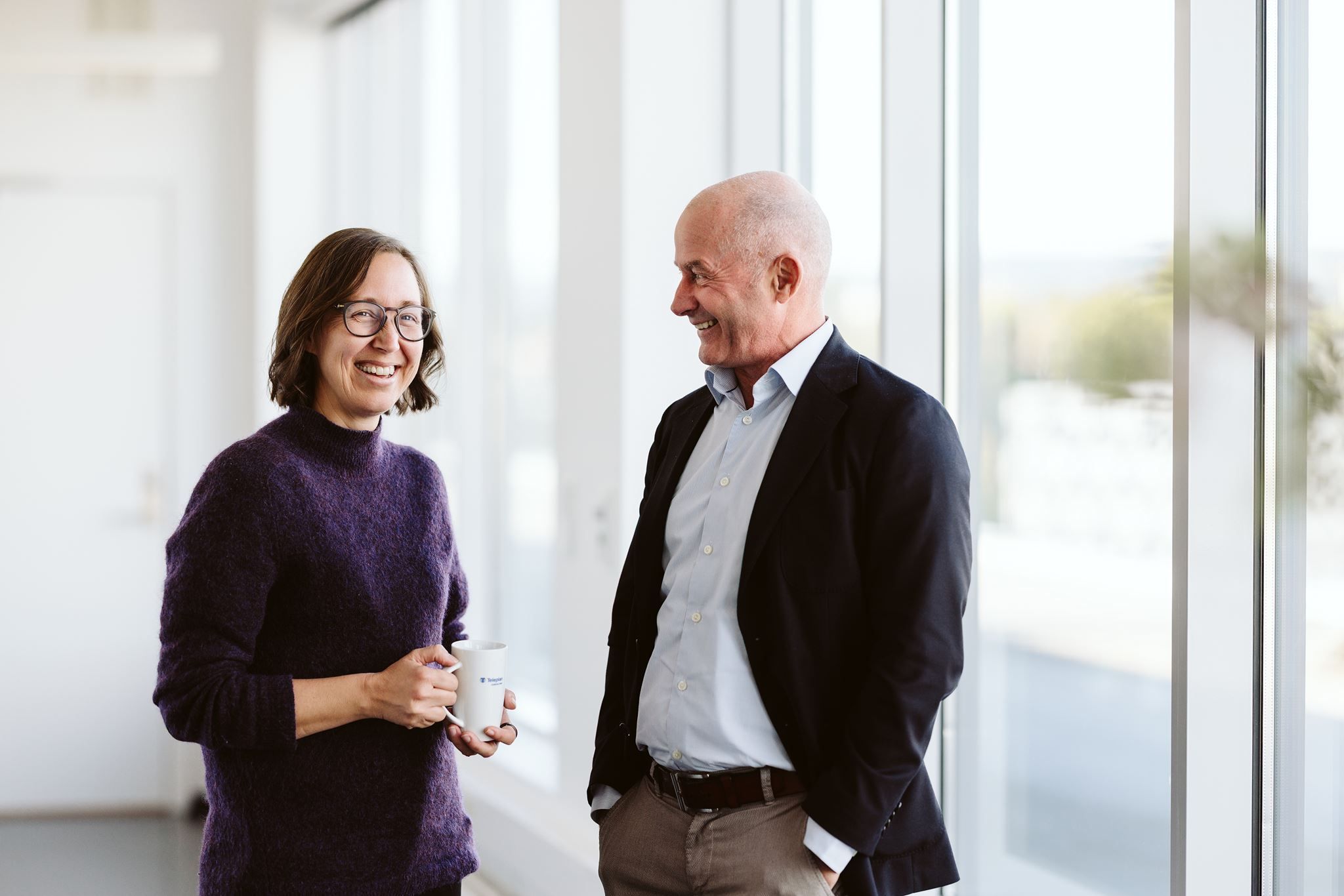 Kvinne med briller og kaffekopp og mann i dress står ved et vindu i et lyst kontor og smiler til hverandreTest