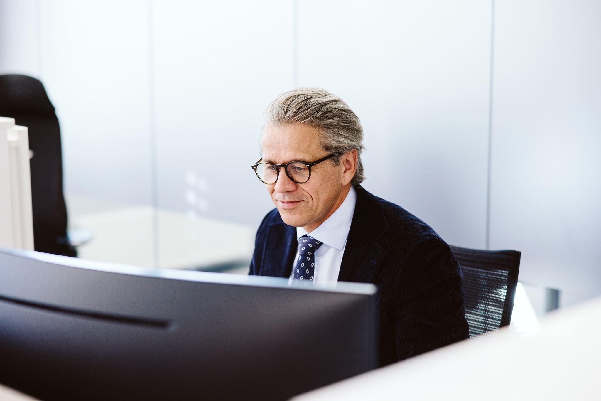Gråhåret mann med briller og dress sitter ved en dataskjerm og jobber i et lyst kontor